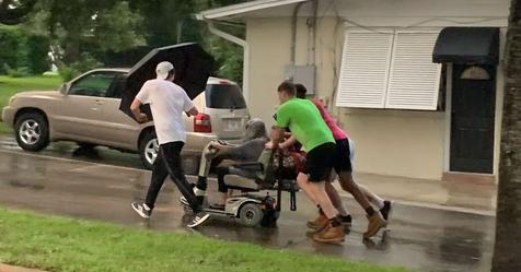 Männer schieben ältere Frau auf kaputten Rollstuhl im strömendem Regen nach Hause