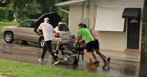 Männer schieben ältere Frau auf kaputten Rollstuhl im strömendem Regen nach Hause